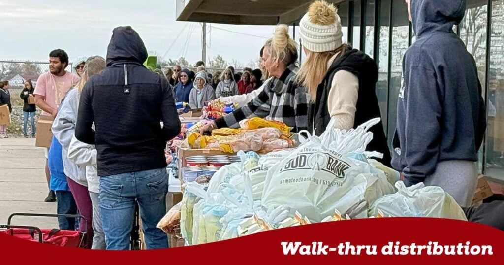 On the right hand side of the image, volunteers stand on one side of a line of tables laden with an assortment of food for distribution. on the left hand side of the table, neighbors are in line getting food from the table. at the bottom of the image is a red banner with the words Walk-thru distribution.